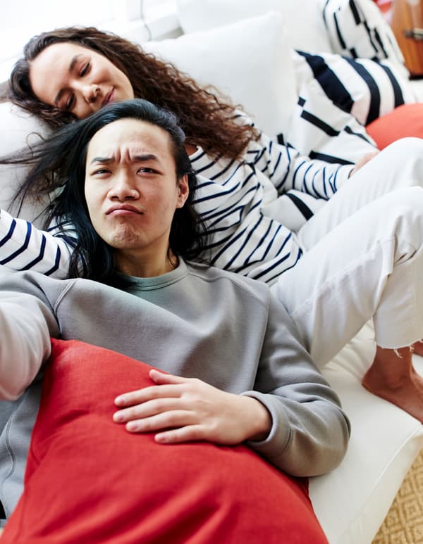 Seorang lelaki dan wanita berbaring berdampingan di atas sofa berwarna putih, dikelilingi oleh kusyen berwarna dan berjalur-jalur hitam putih.