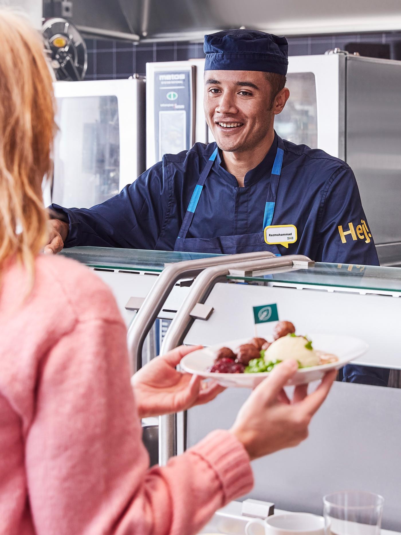 Seorang pelanggan memakai baju panas berwarna merah jambu menerima hidangannya daripada rakan sekerja Bistro Sweden yang sedang tersenyum memakai uniform berwarna biru tua.