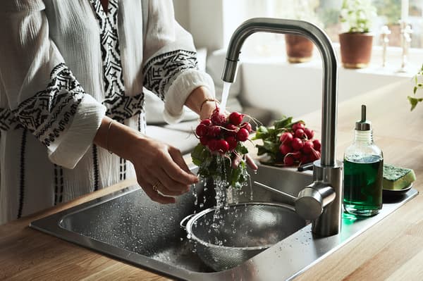 Seorang wanita berdiri di sink membilas lobak menggunakan paip dapur ÄLMAREN.