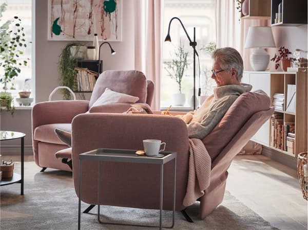 Seorang wanita duduk dengan selesa di kerusi boleh rebah EKOLSUND dengan sarung GUNNARED berwarna coklat merah jambu lembut di sebelah meja kopi.