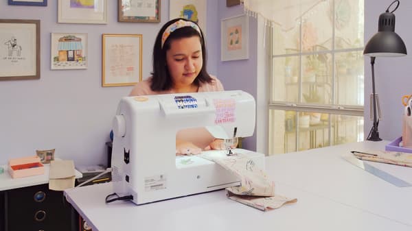 Seorang wanita muda sedang duduk di ruang kecil yang kemas sambil menggunakan mesin jahit untuk membuat sesuatu.