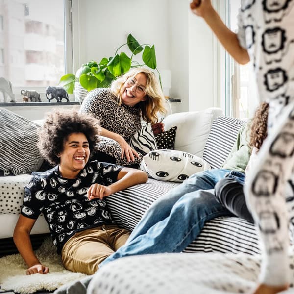 Seorang wanita sedang duduk di sofa dan seorang remaja duduk di atas lantai sedang ketawa sementara seorang lagi kanak-kanak melompat di sofa.
