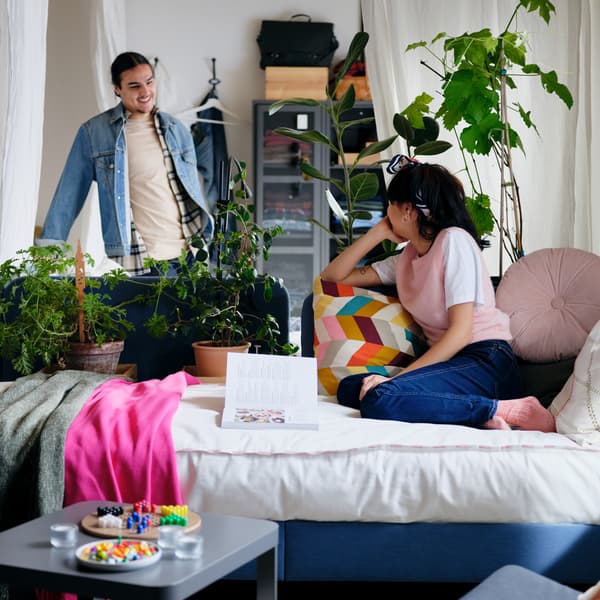 Seorang wanita sedang duduk pada bingkai katil upholsteri BLÅKULLEN dengan kusyen berselerak sedang melihat seorang lelaki yang berdiri di sebelah kabinet IVAR.