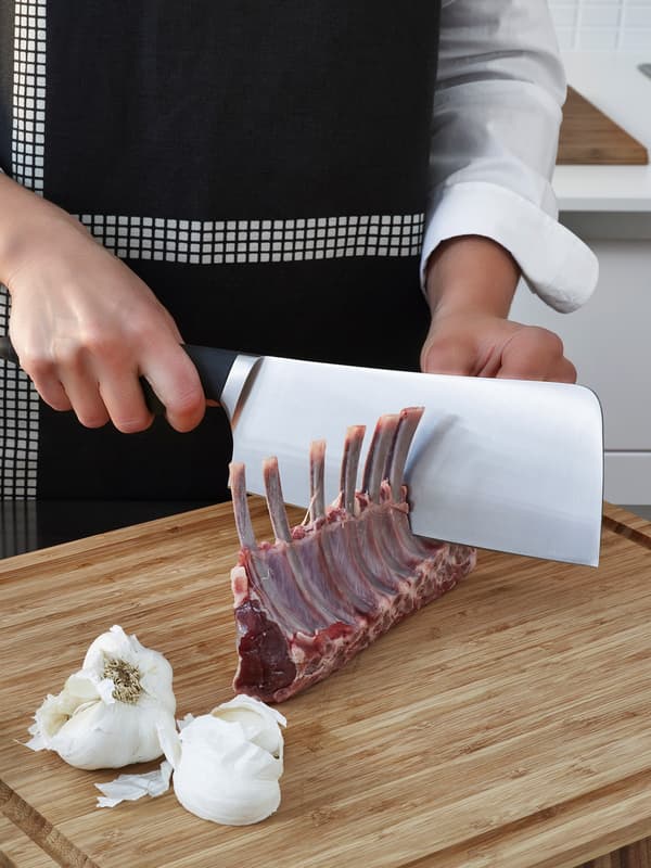 Seorang yang memakai apron berwarna hitam dan putih sedang menggunakan pisau mata lebar dengan pemegang berwarna hitam untuk memotong daging di atas landas cencang kayu.