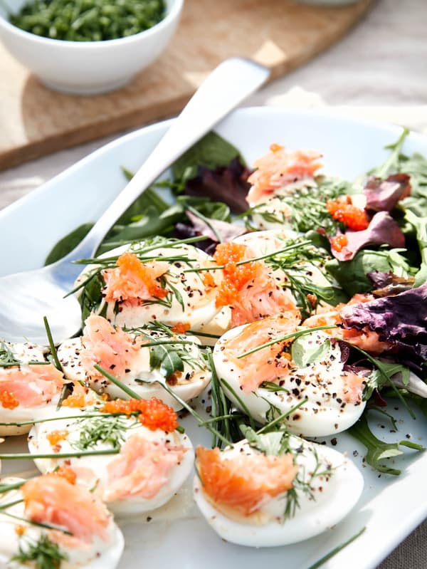 Servírovací tanier IKEA 365+ s polovicami varených vajíčok ozdobenými údeným lososom SJÖRAPPORT, pažítkou a kôprom, v pozadí je miska s nakrájanou pažítkou.