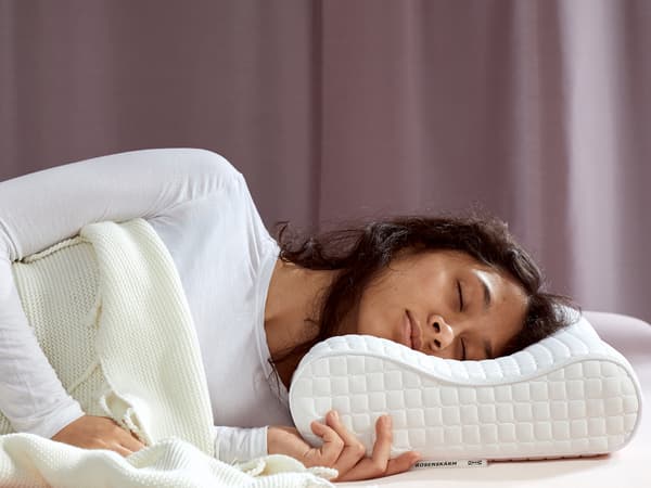 Seseorang berbaju warna putih sedang tertidur di atas bantal ROSENSKÄRM di atas katil dengan langsir berwarna ungu lembut di belakangnya.