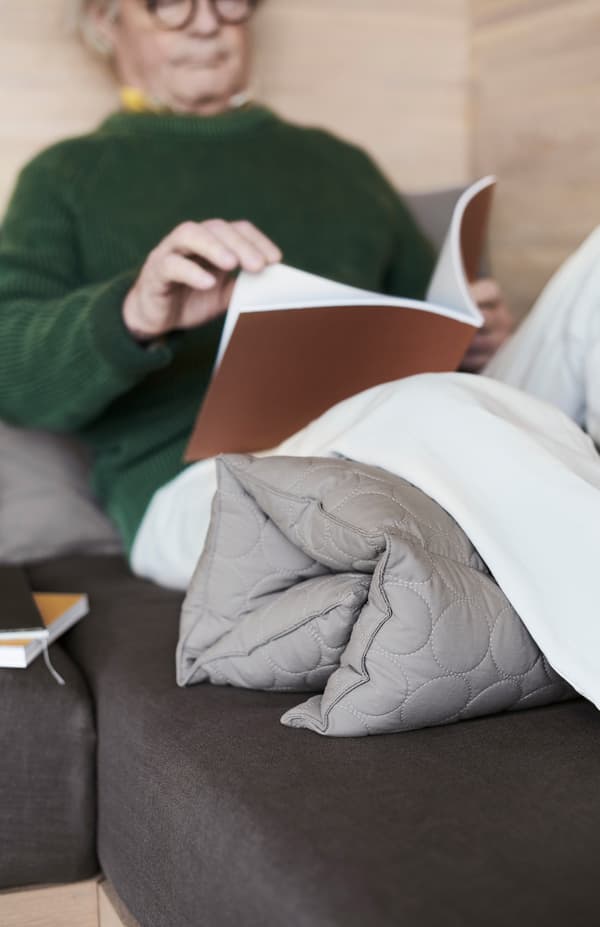 Šesťdesiatnik sa opiera o stenu a číta s multifunkčným vankúšom OMTÄNKSAM zloženým pod kolenami.