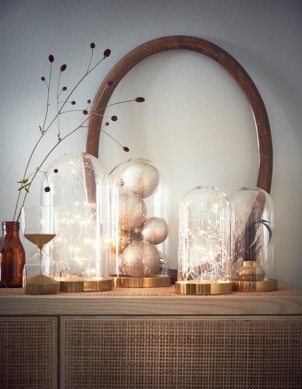 Several decorative glass domes filled with battery powered light chains, baubles and a plant on a cabinet.