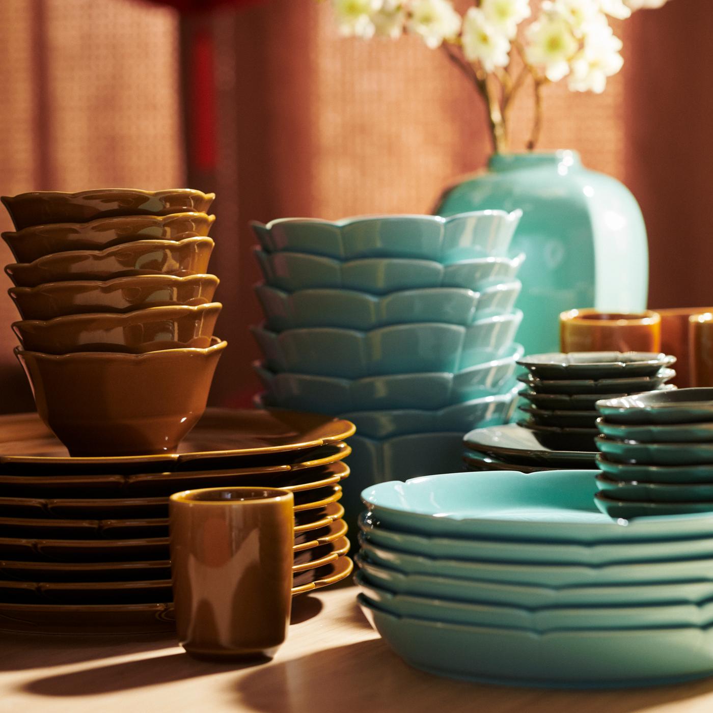 Several FÖSSTA mugs and stacks of FÖSSTA plates and bowls in shades of yellow and turquoise in front of a FÖSSTA vase.