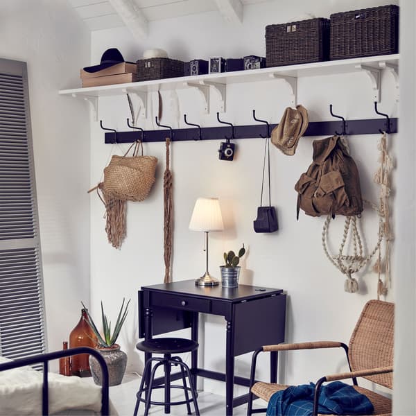 Several IKEA BERGSHULT white wall shelves and PINNIG black racks used to store jackets, bags and accessories near a small table.