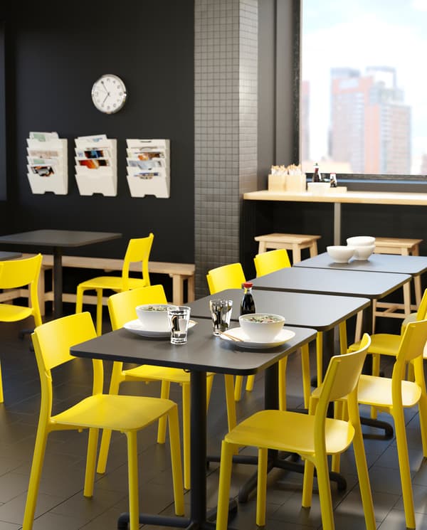 Several STENSELE tables with yellow JANINGE chairs put together in a lunchroom, two white plates and glasses with water.