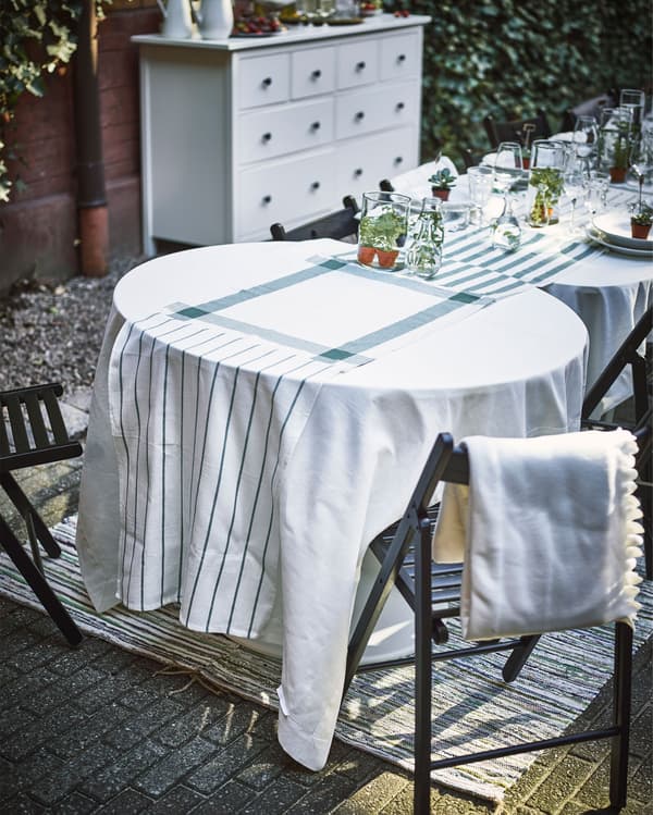 Several tables of different shapes and sizes sit in a row, with white tablecloths and a table runner made of white and green dish towels.