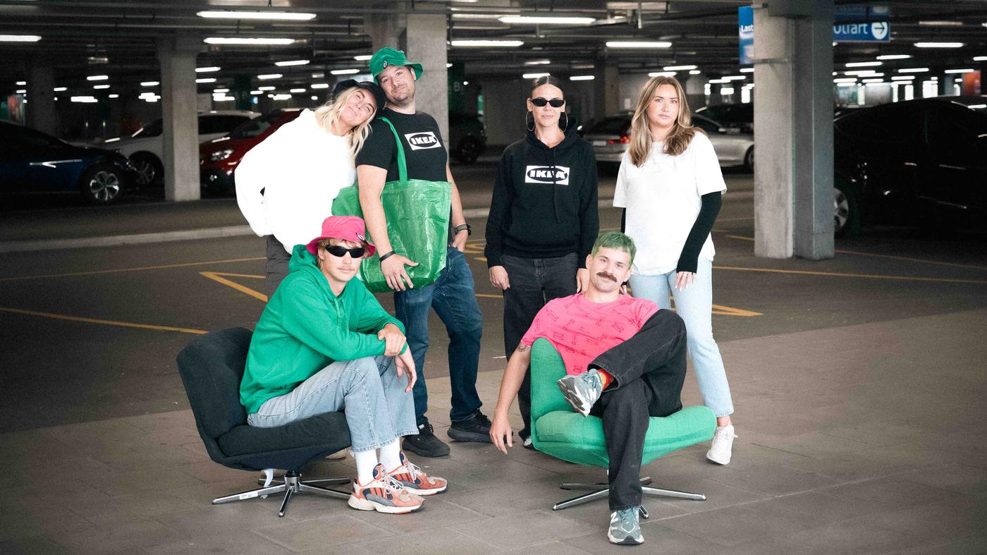 Sex personer befinner sig i ett parkeringsgarage. Fyra står och två sitter i fåtöljer. De har alla kläder på sig som visar IKEA-logotypen. 
