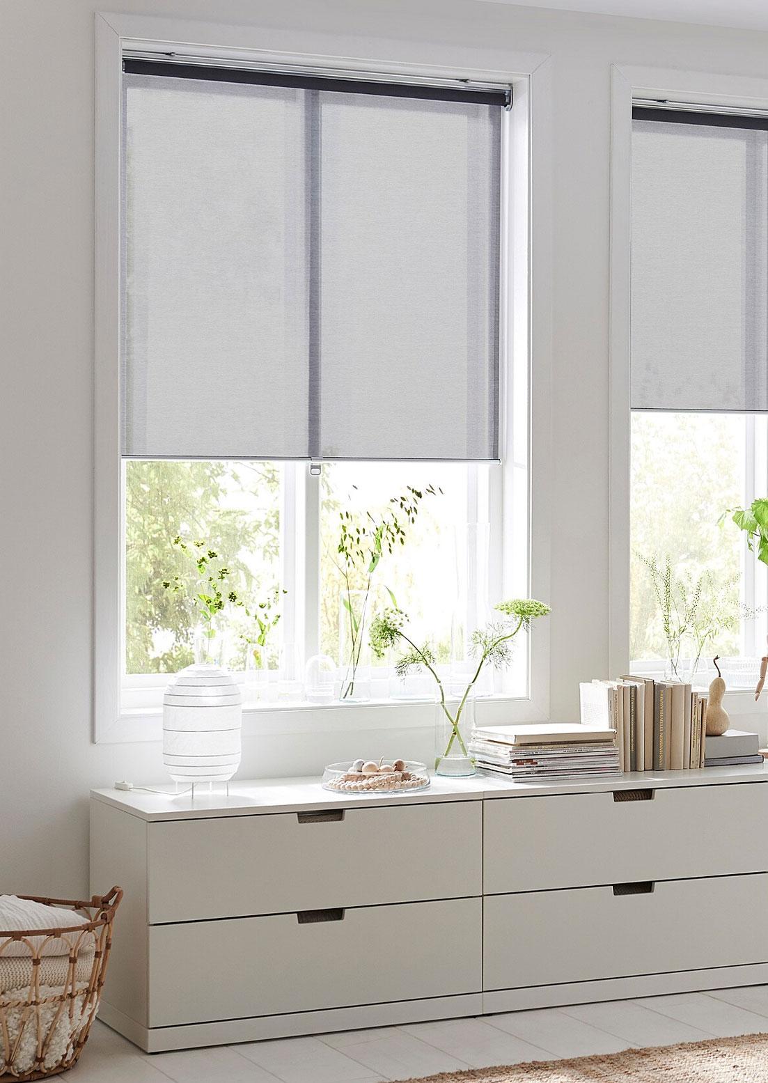 Shelves with books are mounted on the wall beside a window with a FRIDANS block-out roller blind and thin curtains.