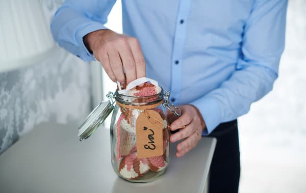 Shirt-clad man placing a crocheted cloth in a KORKEN glass jar. Round the top of the jar is a string with a hand-written tag.