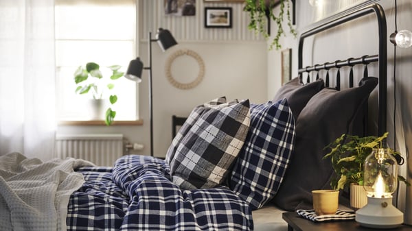 Side profile of a black metal frame bed featuring the TORNTUSS flannel duvet cover, a cream blanket near the foot of the bed, and three sets of pillows in varying patterns and sizes. At the side of the bed is a bedside table with a lamp, coffee cup, and plant. 