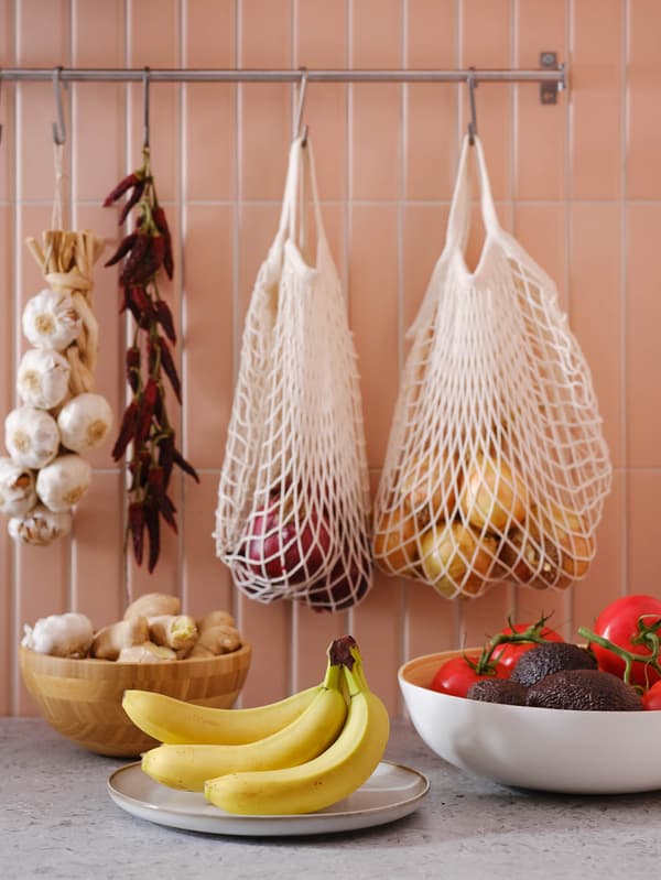 Sieťovky KUNGSFORS s cibuľou visia na tyči s háčikmi KUNGSFORS, pod nimi je misa BLANDA MATT s čerstvým zázvorom, tanier GLADELIG s banánmi a misa RUNDLIG s paradajkami a avokádami.