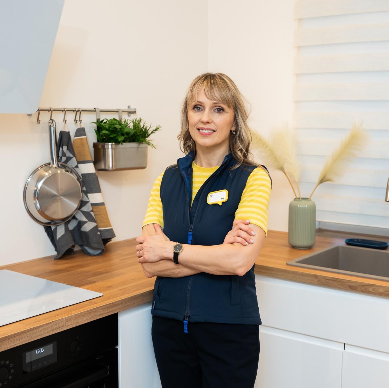 Sieviete IKEA formastērpā stāv gaišā, modernā virtuvē ar sakrustotām rokām; fonā redzama koka darba virsma, balti skapji, izlietne un pie sienas piekārti virtuves piederumi.