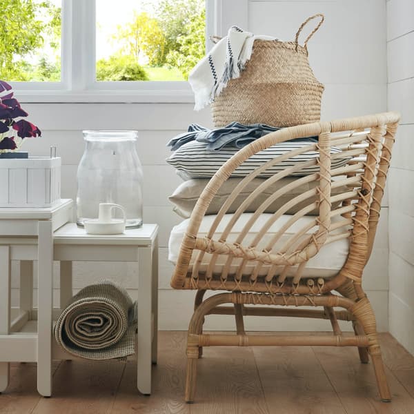Silla de fibras naturales BUSKBO color cafe claro o beige. Tiene encima dos coijnes, de los cuales uno es rayado y el otro es de color gris claro. Sobre ellos hay una cesta de fibras también.