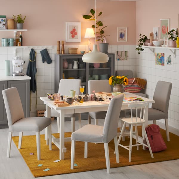 Sillas con funda de la serie BERGMUND, con un comedor con flores y decoración sobre el, lamparas y adornos colgados en el techo.