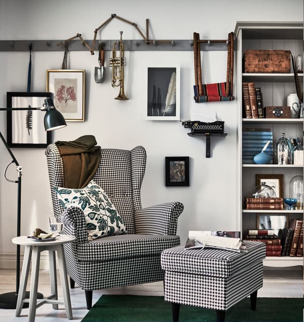 Sillón de IKEA orejero de cuadros vichy con un reposapiés del mismo estampado, una mesa auxiliar gris de de madera redonda y una lámpara de pie negra. Es de estilo escandinavo tradicional de IKEA.
