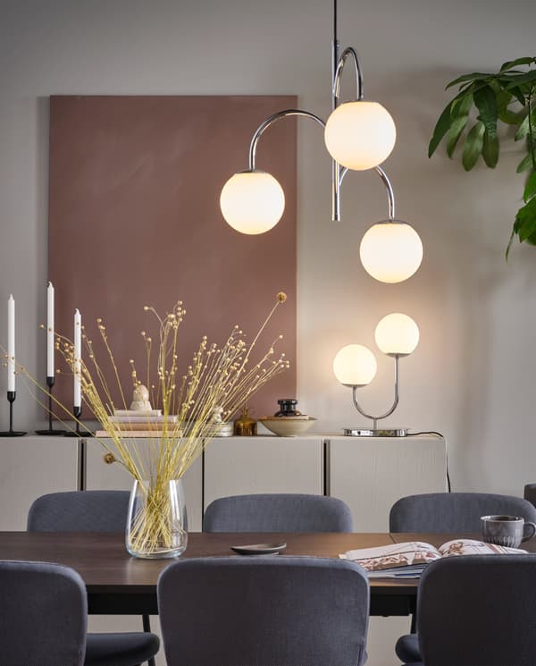 SIMRISHAMN 3-armed chrome-plated pendant lamp hangs over a dining table, and SIMRISHAMN table lamp stands on a cabinet.