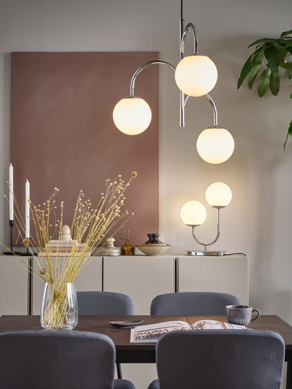 SIMRISHAMN 3-armed chrome-plated pendant lamp hangs over a dining table, and SIMRISHAMN table lamp stands on a cabinet.