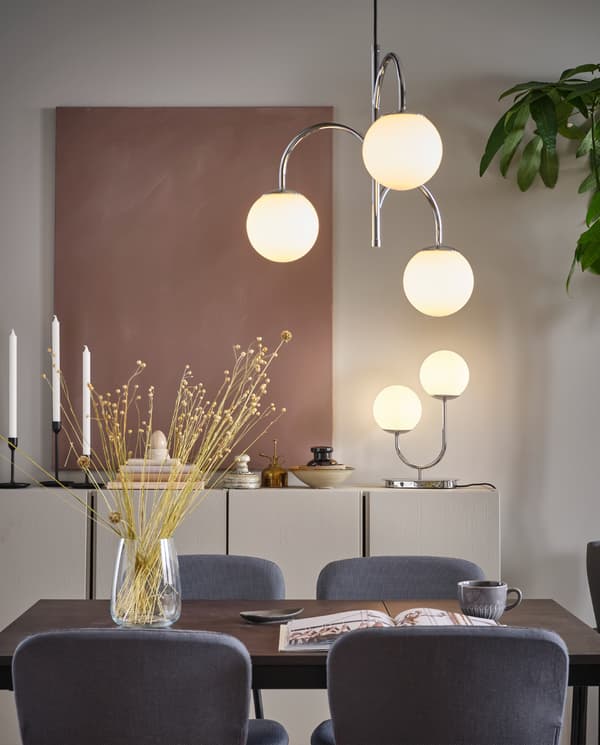 SIMRISHAMN chromen hanglamp met 3 armen boven een eettafel, met een SIMRISHAMN tafellamp op een kastje.