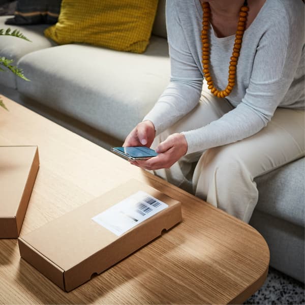 Sitting comfortably on a modern gray IKEA sofa with a green plant next to it, a person wearing a gray top and cream-colored pants scans a package barcode with their smartphone on a light-colored wooden IKEA coffee table.