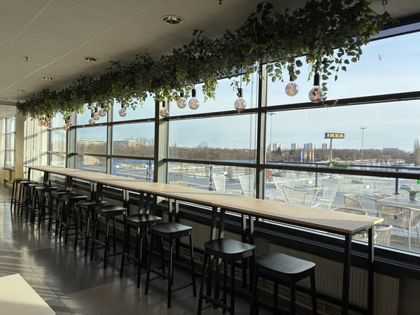 Sittplatser vid fönster i IKEA Malmös personalmatsal med höga bord, pallar, stora glaspartier och hängande växter som släpper in dagsljus.