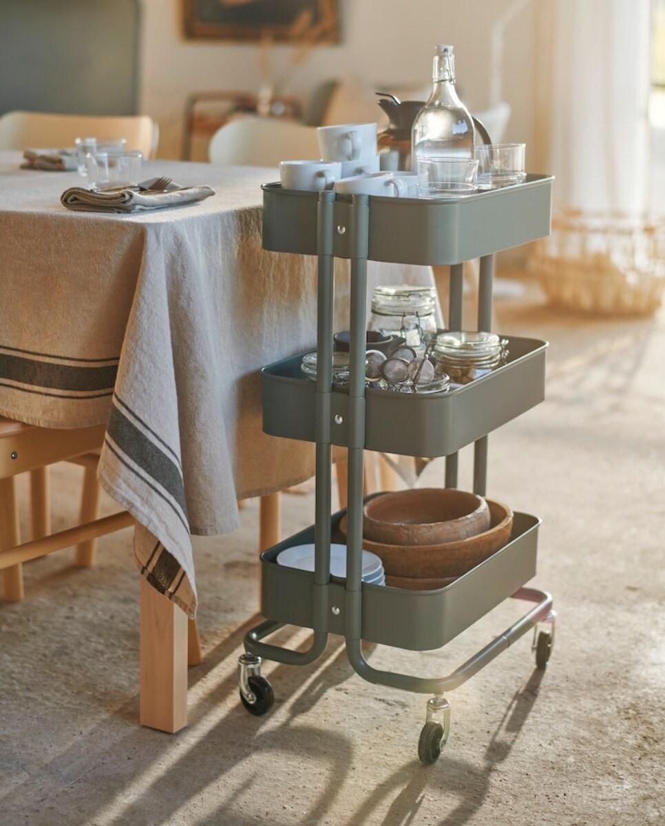Siva kolica s tri police, na kojima su čaše, zdjele i boca, stoje pored stola prekrivenog stolnjakom u blagovaonici.