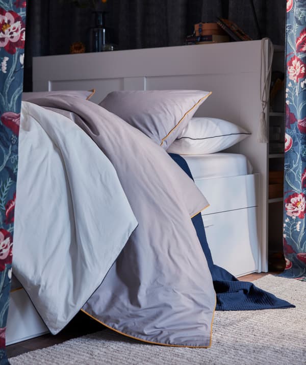 Sivá postená obliečka IKEA KUNGSBLOMMA s žltými okrajmi na posteli s nadýchanými textíliami.