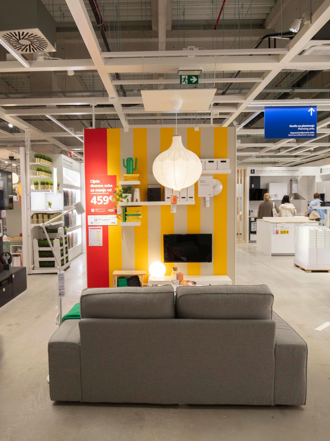 Siva sofa ispred zida s žutim i bijelim prugama, policama, TV-om i visećom lampom. Na zidu je crveni plakat s cijenom 459 kn, u pozadini se vidi trgovina namještajem.