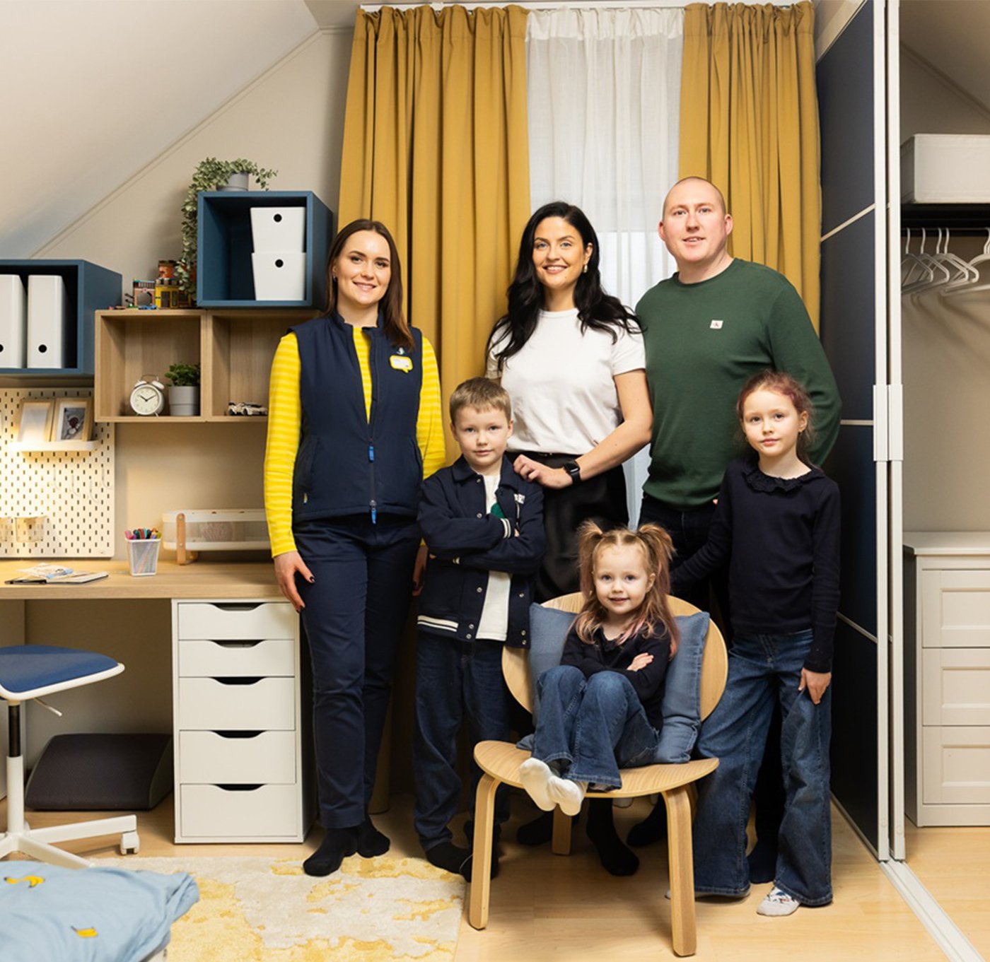 Six people are standing together in the renovated children’s room after the makeover. Two adults and three children are posing in the finished interior featuring coordinated furniture, storage systems, and a study area; on the left stands an IKEA designer.