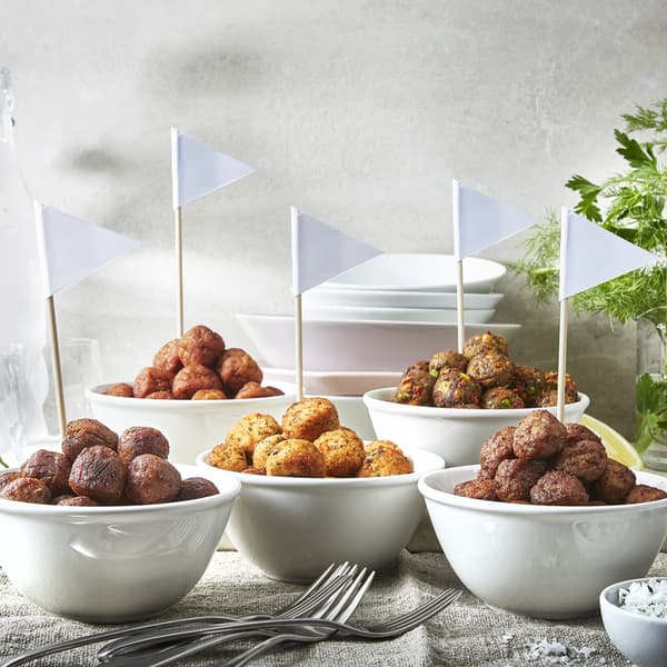 Six white bowls of assorted meatballs with flags, forks in front, herbs and stacked plates in background on cloth surface.
