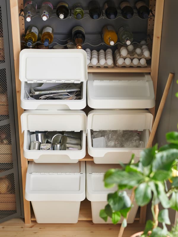 Six white SORTERA waste sorting bins with lids, arranged in two stacks of three, in a wooden stacking rack beneath eggs and bottles