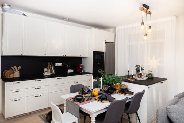 Skats uz virtuves darba zonu ar plīti, cepeškrāsni un sadzīves tehniku; blakus atrodas ēdamgalds un logs ar aizkariem.