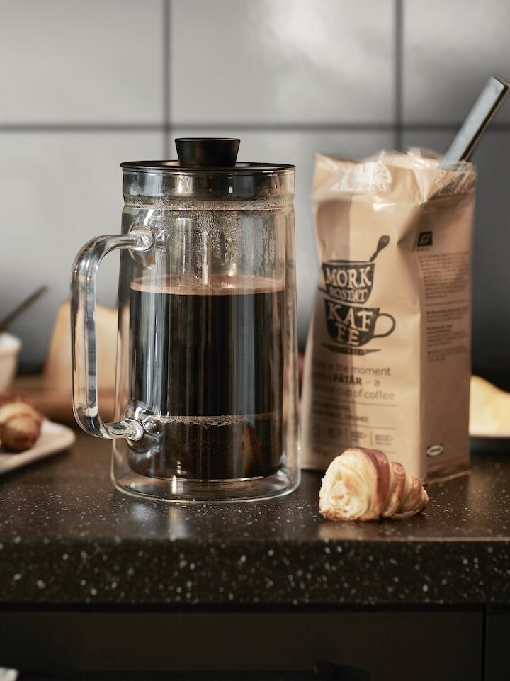 Sklenený french press s čiernou kávou stojí na kuchynskej linke vedľa otvoreného balíčka mletej kávy a malého croissantu. V pozadí sú dlaždice a ďalšie pečivo.