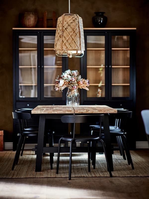 SKOGSTA dining table with a flower vase under a KNIXHULT pendant, surrounded by dark chairs. Dark cabinets by the far wall.
