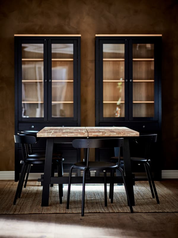 SKOGSTA dining table, with light-wood top and dark legs, surrounded by anthracite YNGVAR chairs. Dark cabinets by the wall.