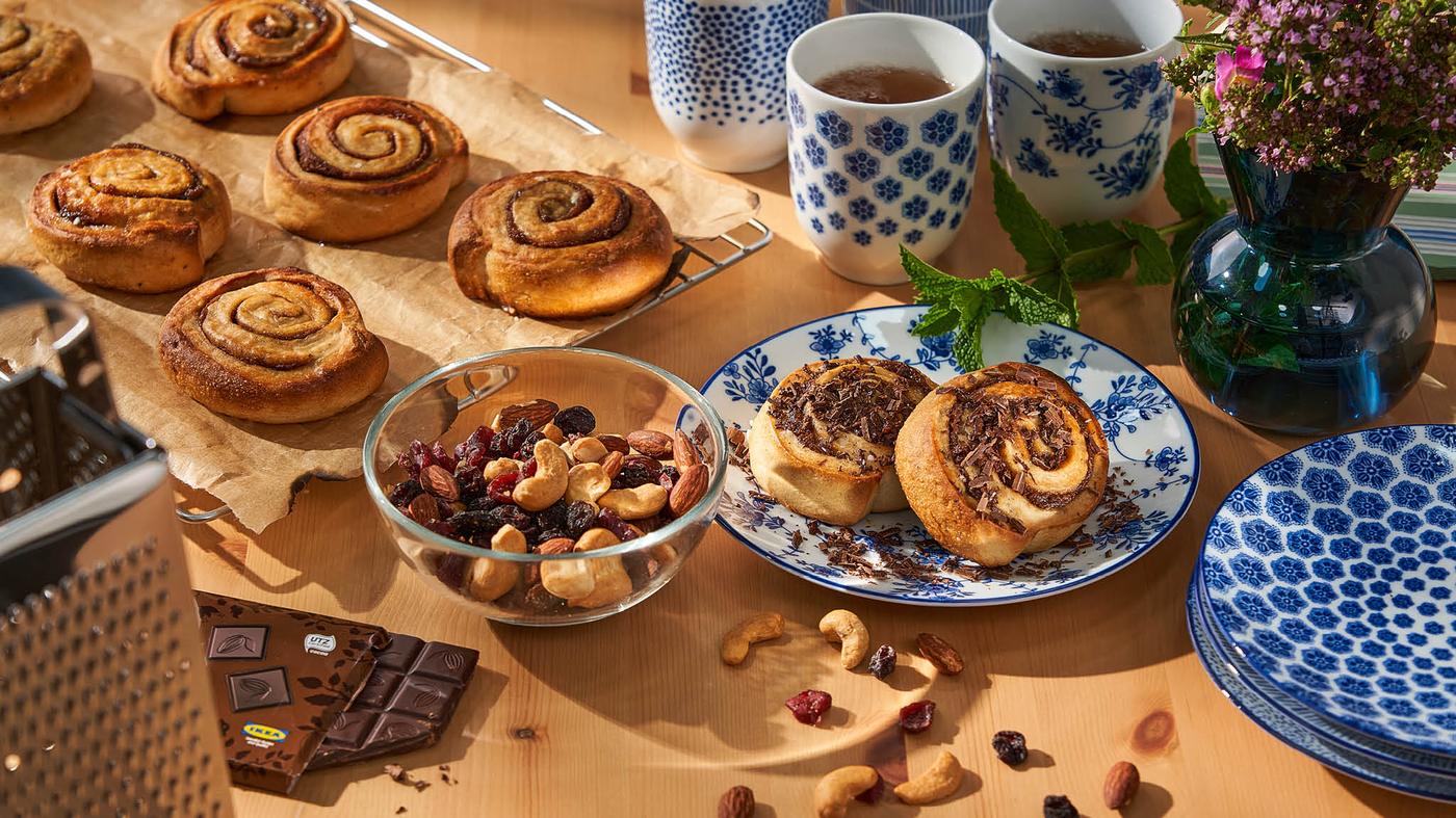Škoricové koláčiky KAFFEREP na chladiacej mriežke LÄTTBAKAD, pred nimi je miska BLANDA s orieškami MUNSBIT, dezertné taniere ENTUSIASM s ďalšími dvomi koláčikmi s nastrúhanou čokoládou, hrnčeky ENTUSIASM s čajom a váza/svietnik STOCKHOLM 2017.
