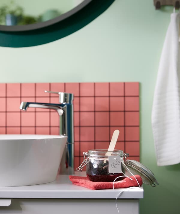 Skrinka s umývadlom v kúpeľni so zloženým uterákom VÅGSJÖN, na ktorom je položená dóza KORKEN s domácim peelingom, označená ručne napísaným štítkom.