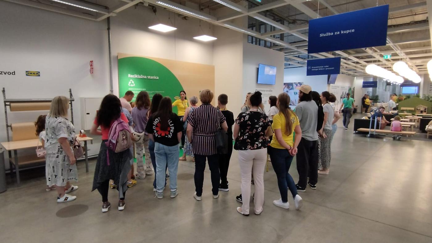 Skupina ljudi okupljena ispred zelenog zida s natpisom "Reciklažna stanica" u trgovini.