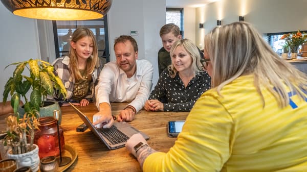 Skupina ljudi sjedi za stolom, gledajući u laptop i mobitel. Na stolu su biljke i svijeće.