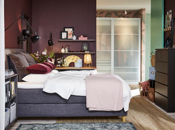 Slaapkamer met een donkergrijs boxspring, zwarte wandlampen, een jute vloerkleed, een zwart-bruine ladekast en donkerrode kussenhoezen