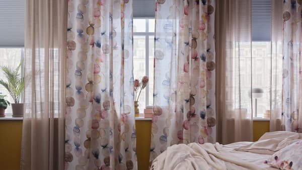 Slaapkamer met een rij ramen met veelkleurige SÄCKMAL gordijnen, beige doorschijnende gordijnen en witte verduisterende plisségordijnen.