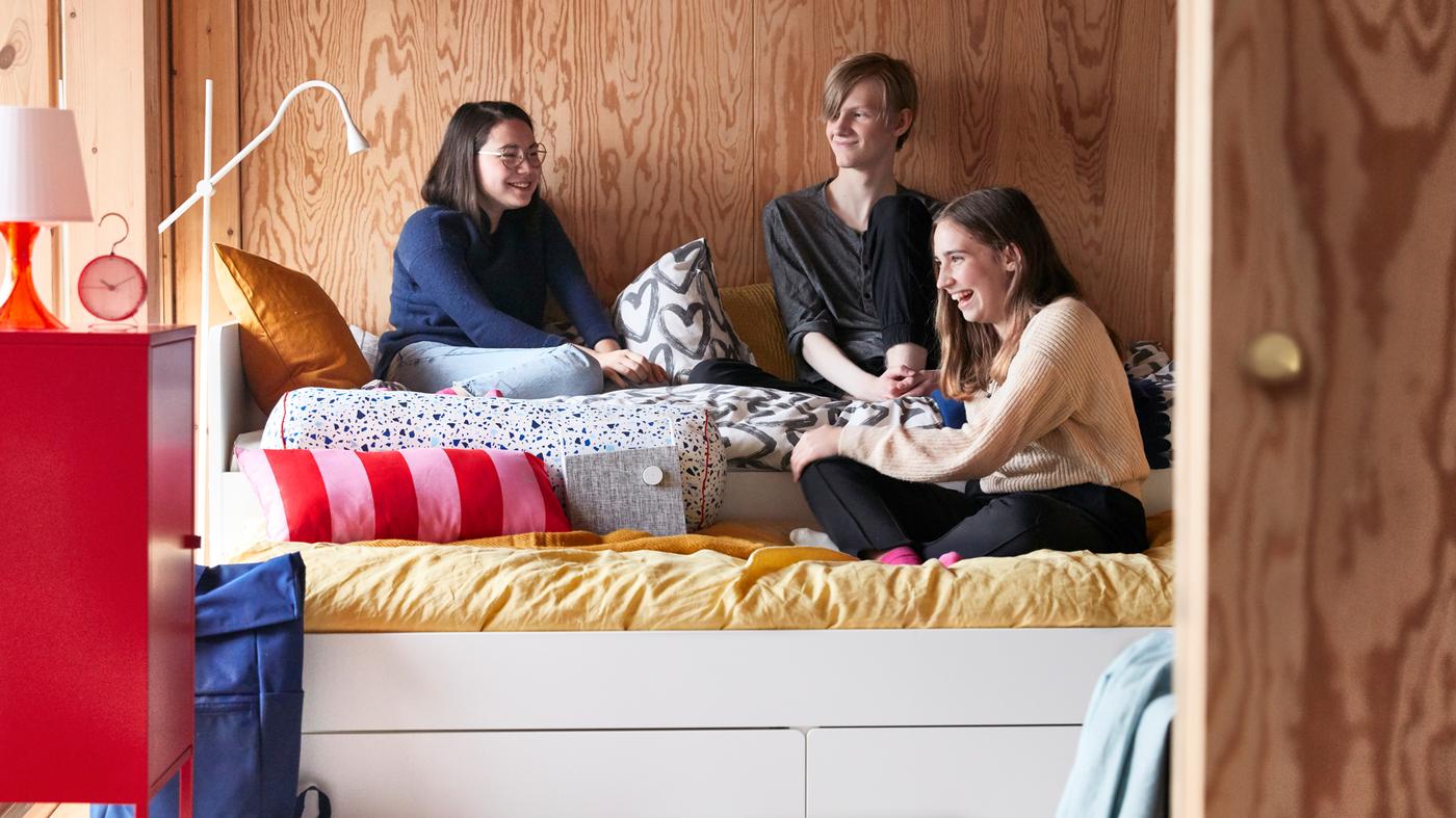화이트 SLÄKT 슬렉트 침대프레임, 다양한 색상의 쿠션과 이불커버로 꾸민 10대 아이 방에서 놀고 있는 세 명의 친구들.