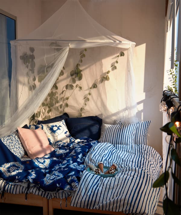 Slnečný balkón s manželskou posteľou s mäkkými vankúšmi a modro-bielymi posteľnými obliečkami, sieťou proti komárom IKEA SOLIG a rastlinami okolo.