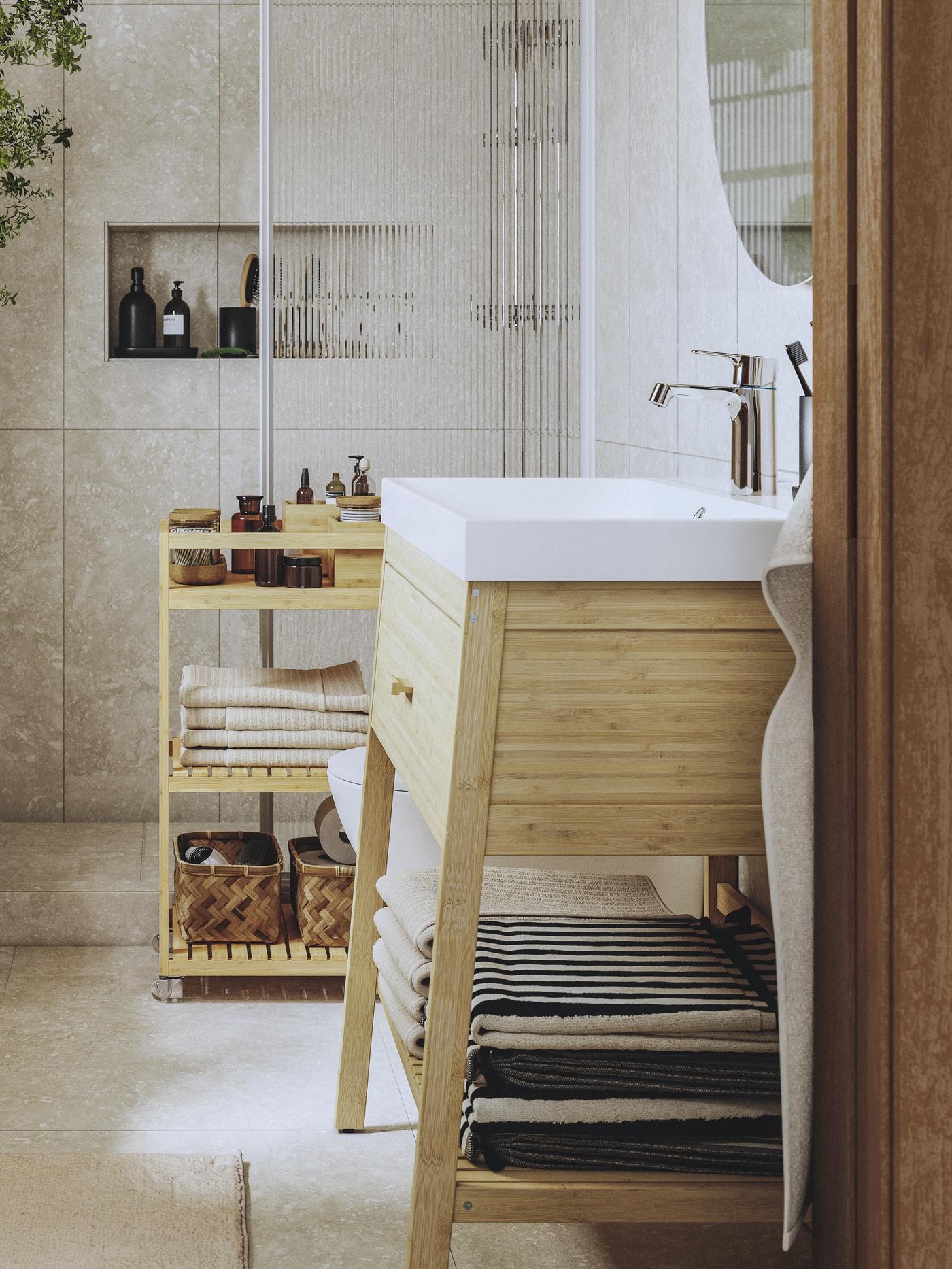 Słoneczna łazienka z wózkiem, krzesłem z wieszakiem i szafką pod umywalkę RÅGRUND wykonanymi z naturalnego bambusa. W kącie stoi duża roślina.