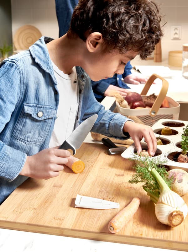 เด็กชายถือมีด SMÅBIT/สมอบิท หั่นเศษผักบนเขียงแล้วใส่ชิ้นที่หั่นลงในดินที่อยู่ในพิมพ์มัฟฟิน VARDAGEN/วาร์ดาเกน